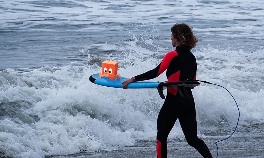 In diesem Bild sieht man eine Surferin mit ihrem Surfbrett unter dem Arm. Auf dem Surfbrett ist eine kleine Pappkrabbe.