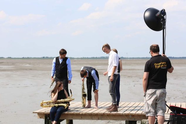 In diesem Bild sieht man 6 Personen die sich für ein Shooting vorbereiten. Im Vordergrund steht eine Person mit einem Fotolicht in der Hand. Die restlichen Personen stehen auf einem Steg in der Mitte des Bildes. Drei von diesen Personen haben Instrumente in der Hand